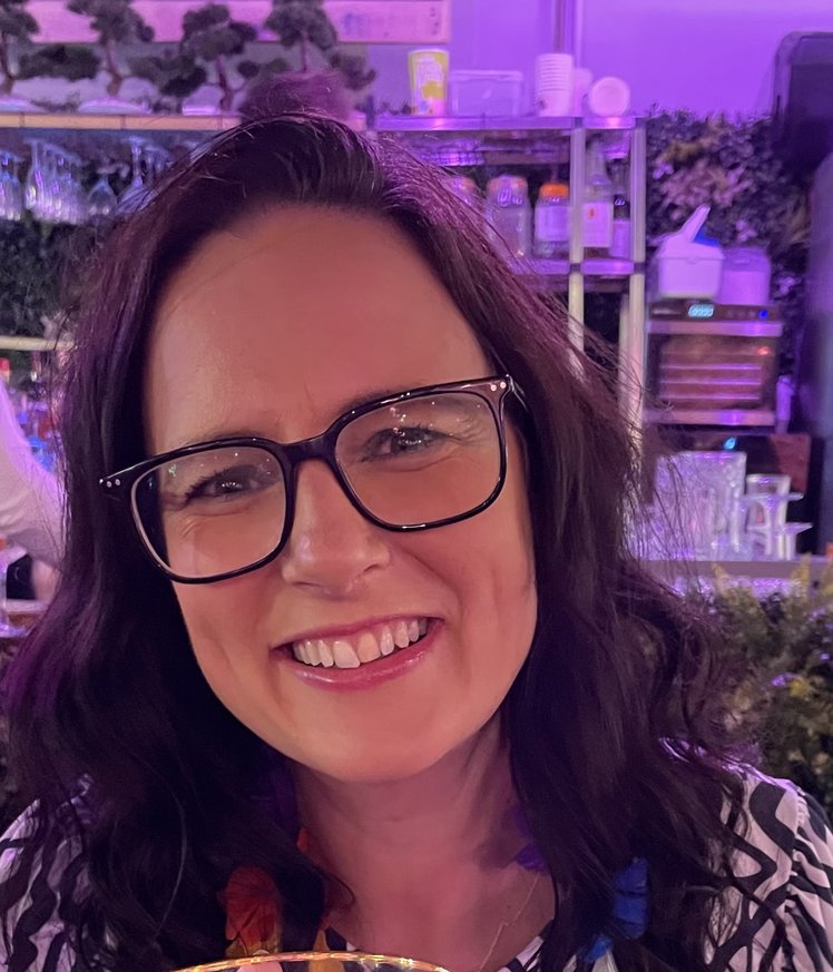 A white woman with long dark hair and glasses smiles into the camera.