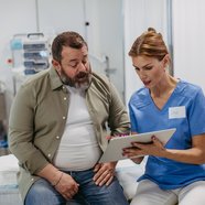 A bearded white man in a white t-shirt and brown shirt talks to a female health professional wearing a name badge and blue scrubs.