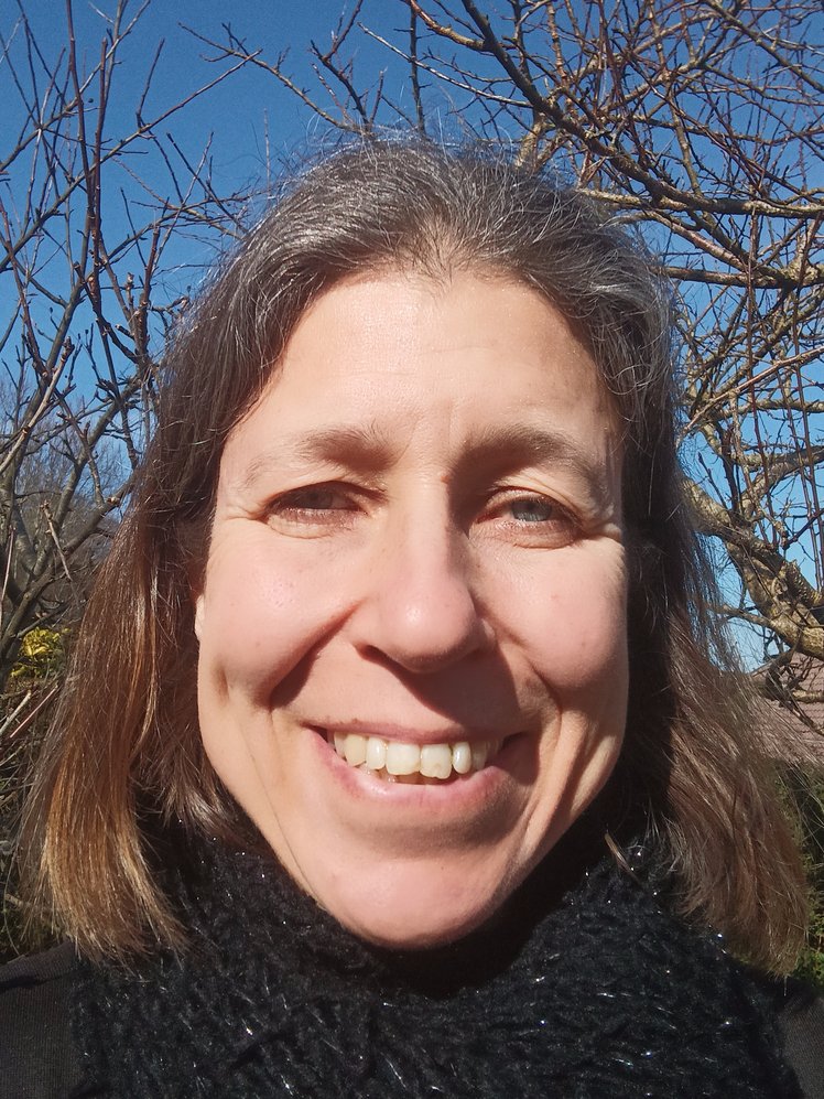 A white woman with brown hair and wrapped in a black scarf smiling with bare winter trees and blue sky behind