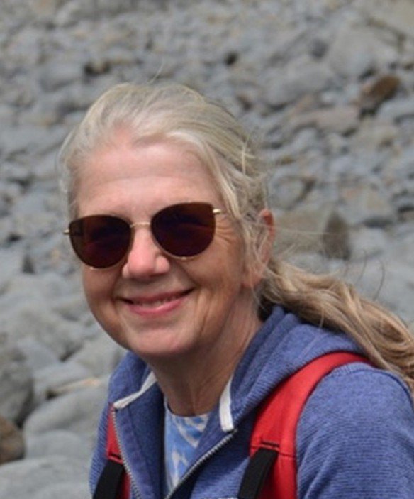 A white woman with long windswept blonde hair in a ponytail smiles at the camera. There is a pebbled beach behind her.