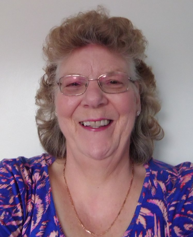 A white woman with wavy strawberry blonde hair smiles cheerily at the camera. She is wearing glasses and a purple and pink patterned top.