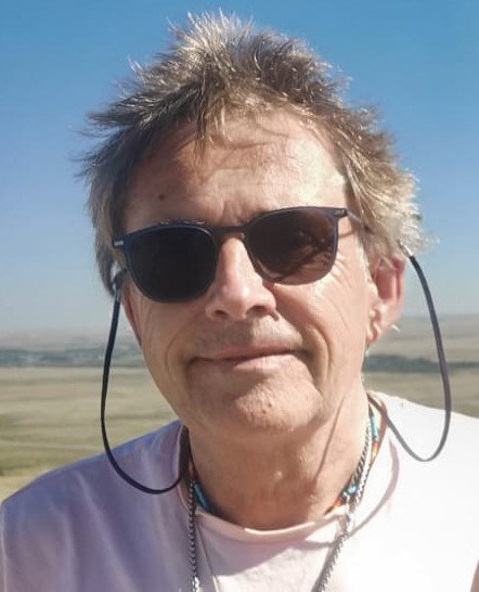 A white man with short greying hair and sunglasses smiles at the camera. There is a beach behind him.